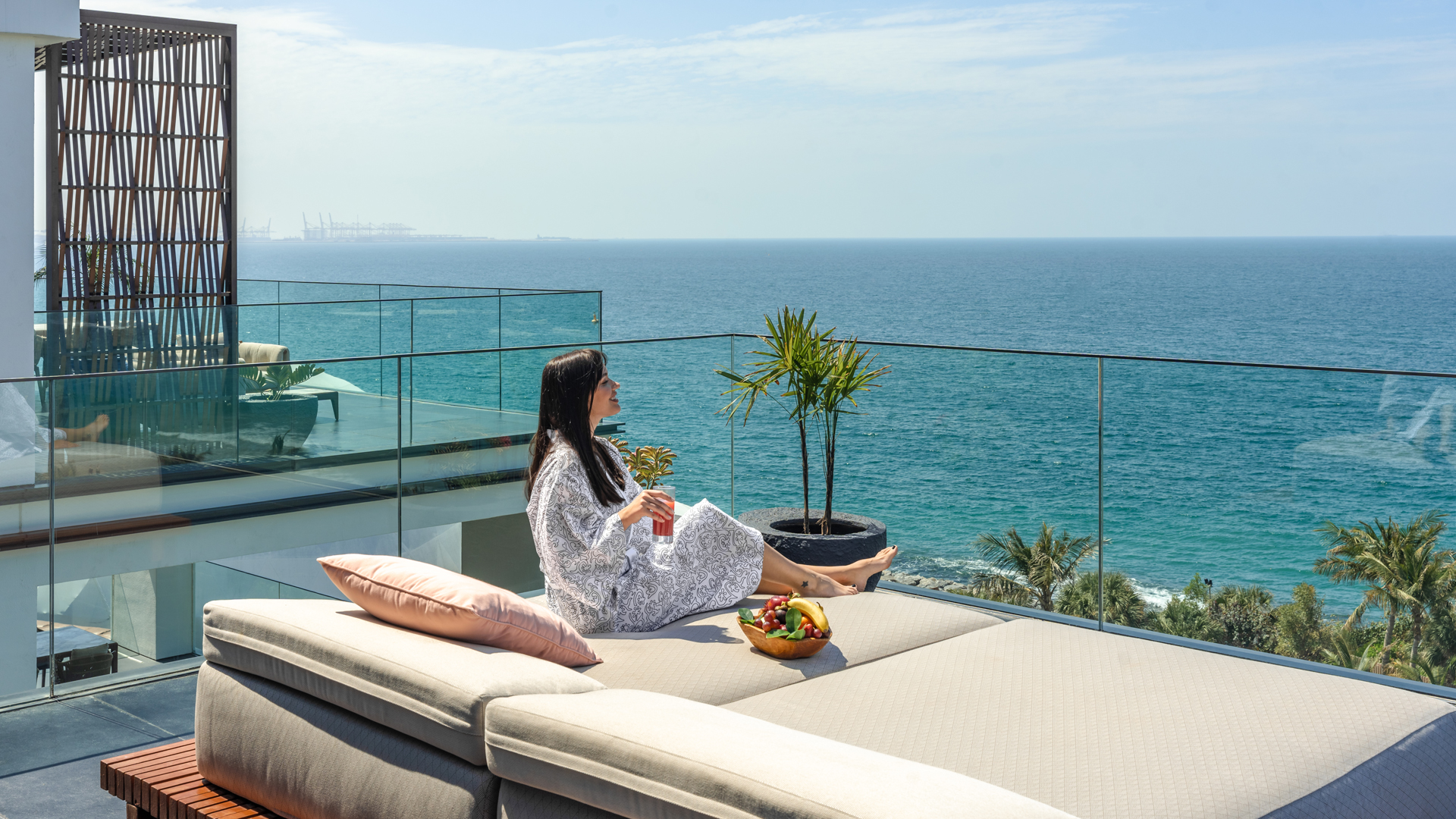 Woman relaxing on a rooftop daybed in a robe, enjoying fresh juice and a fruit platter with panoramic views of the Arabian Gulf at Banyan Tree Dubai.