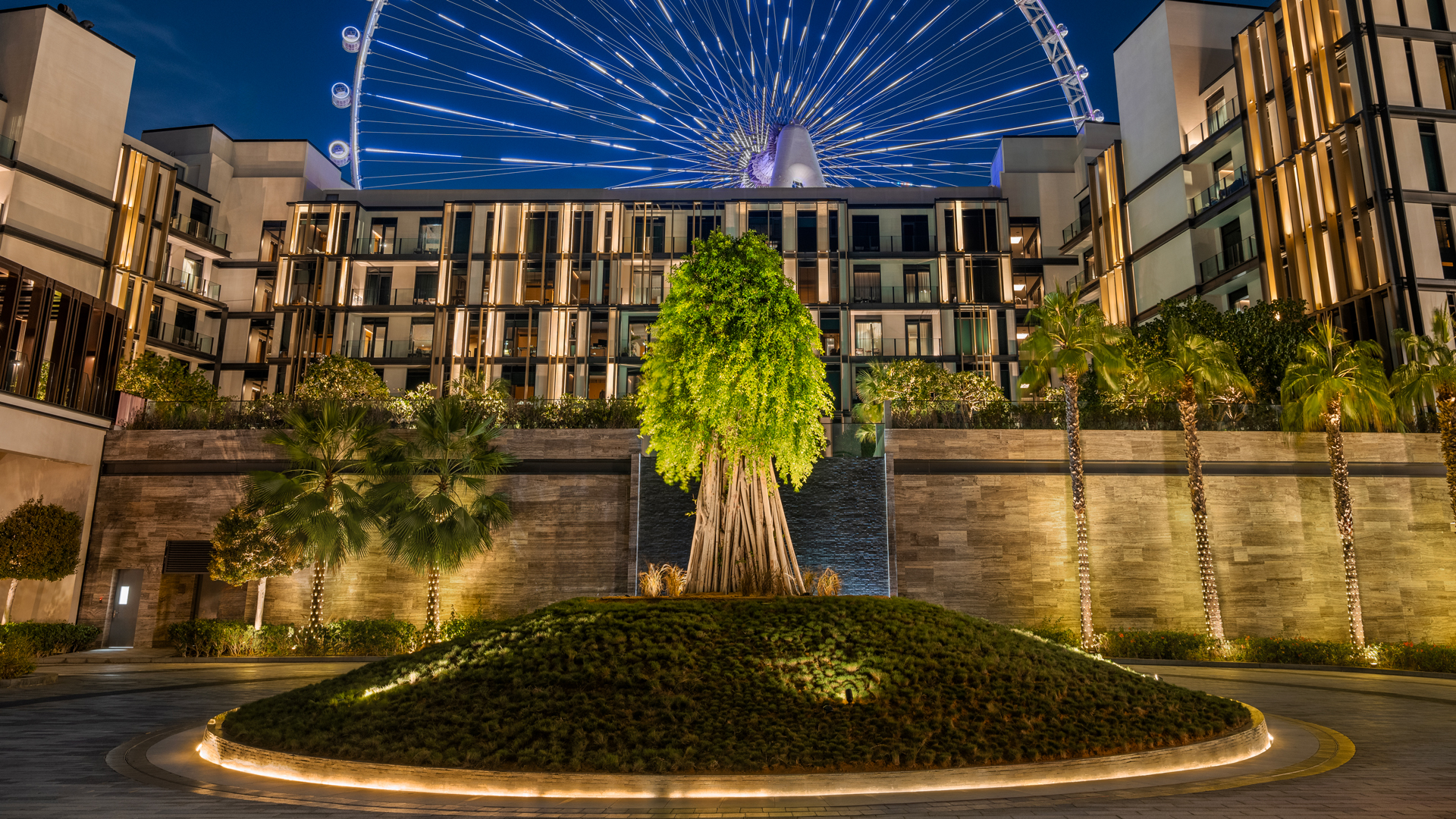 Nighttime view of Banyan Tree Dubai with illuminated banyan tree centerpiece, modern resort façade, and Ain Dubai observation wheel glowing in the backdrop.
