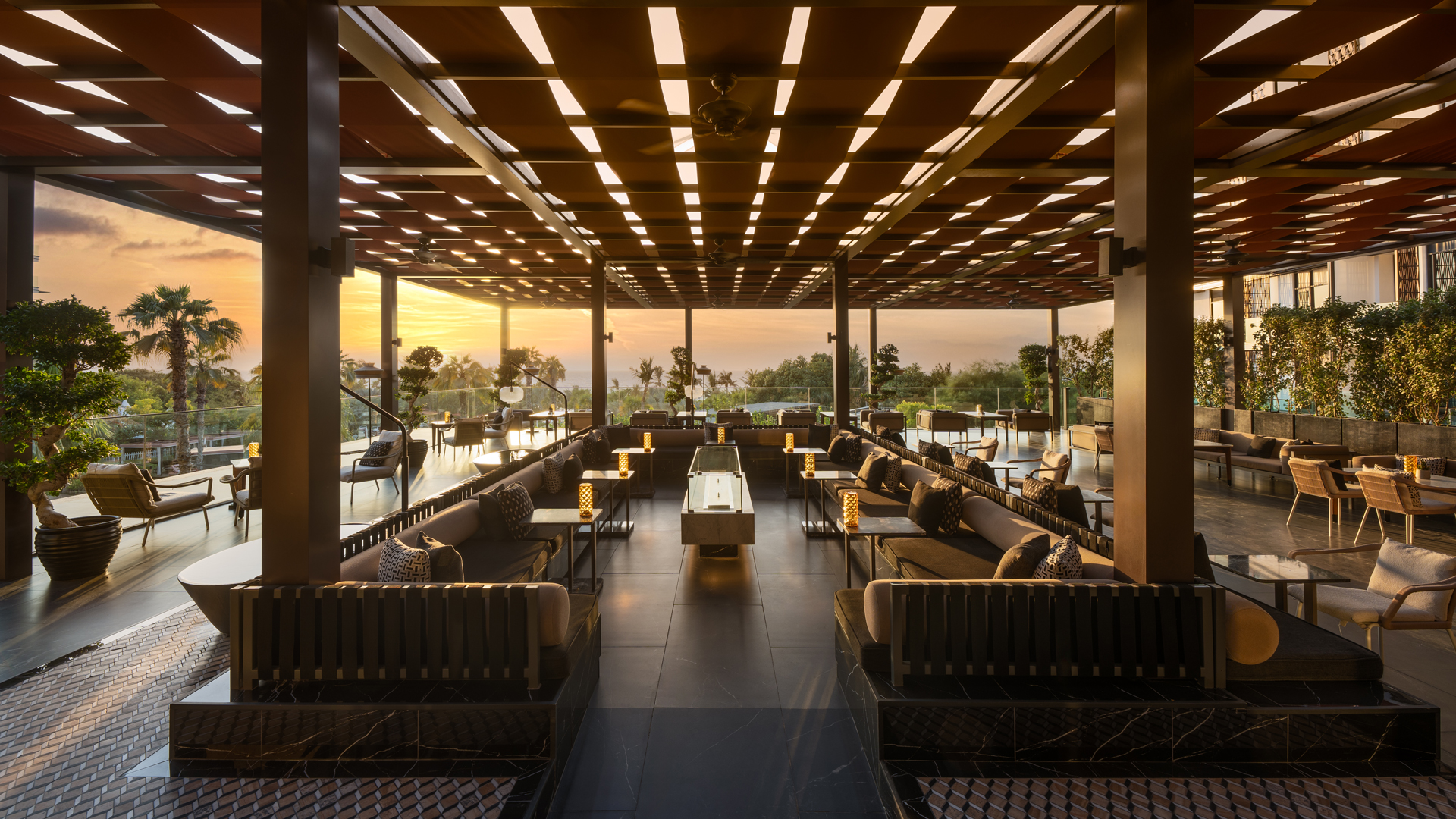 Modern terrace lounge at Tocha overlooking lush greenery and the Arabian Gulf, illuminated by a golden sunset through geometric ceiling panels.