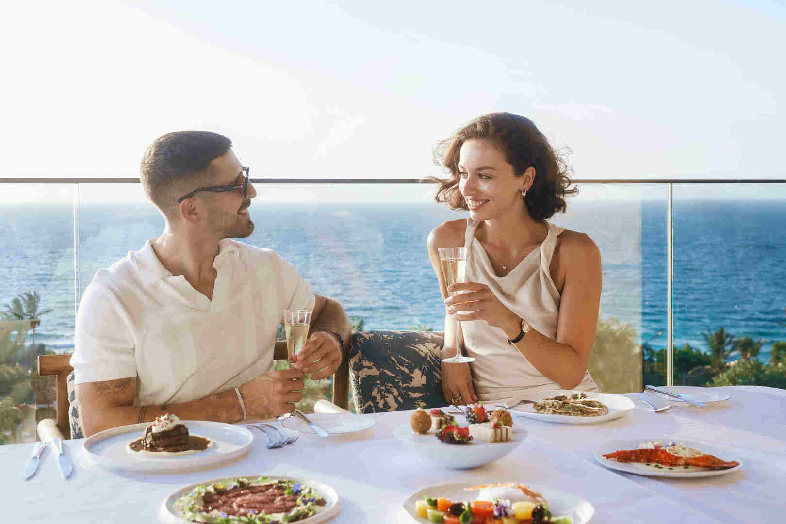 A couple toasts with champagne while enjoying a luxurious private dinner on a terrace with stunning ocean views, surrounded by an elegant setup of gourmet dishes.