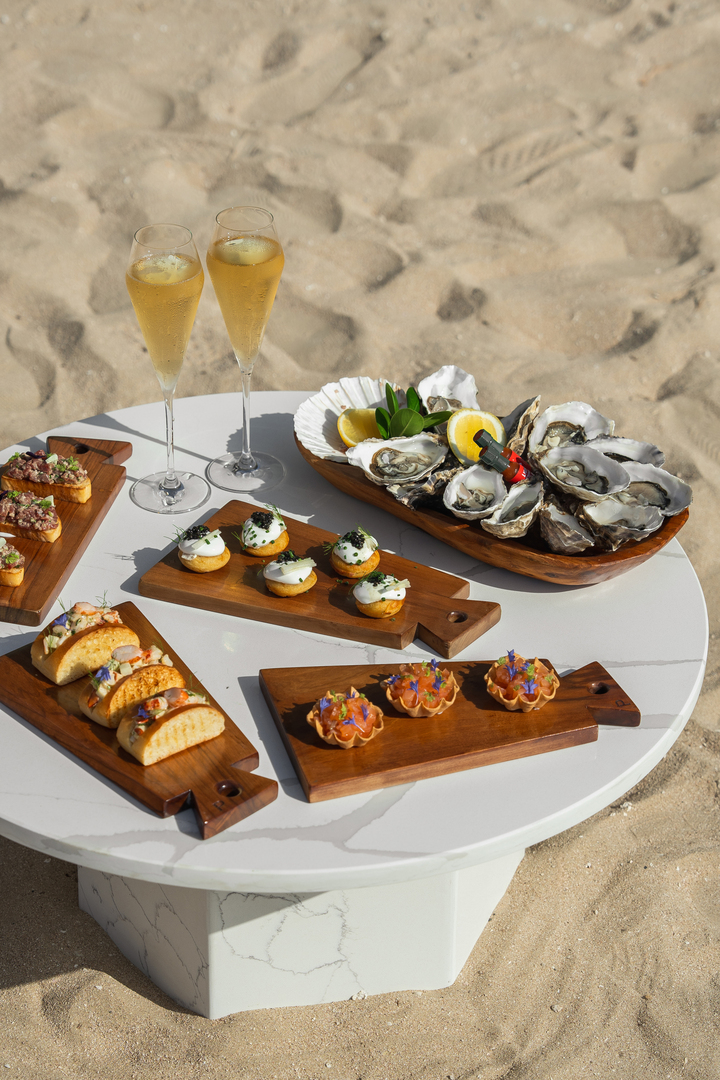bt-dubai-resort-experiences-flavours-by-the-shore A beachfront table set with gourmet bites including oysters, canapés, and mini tacos, complemented by two glasses of champagne, offering a luxurious seaside dining experience.