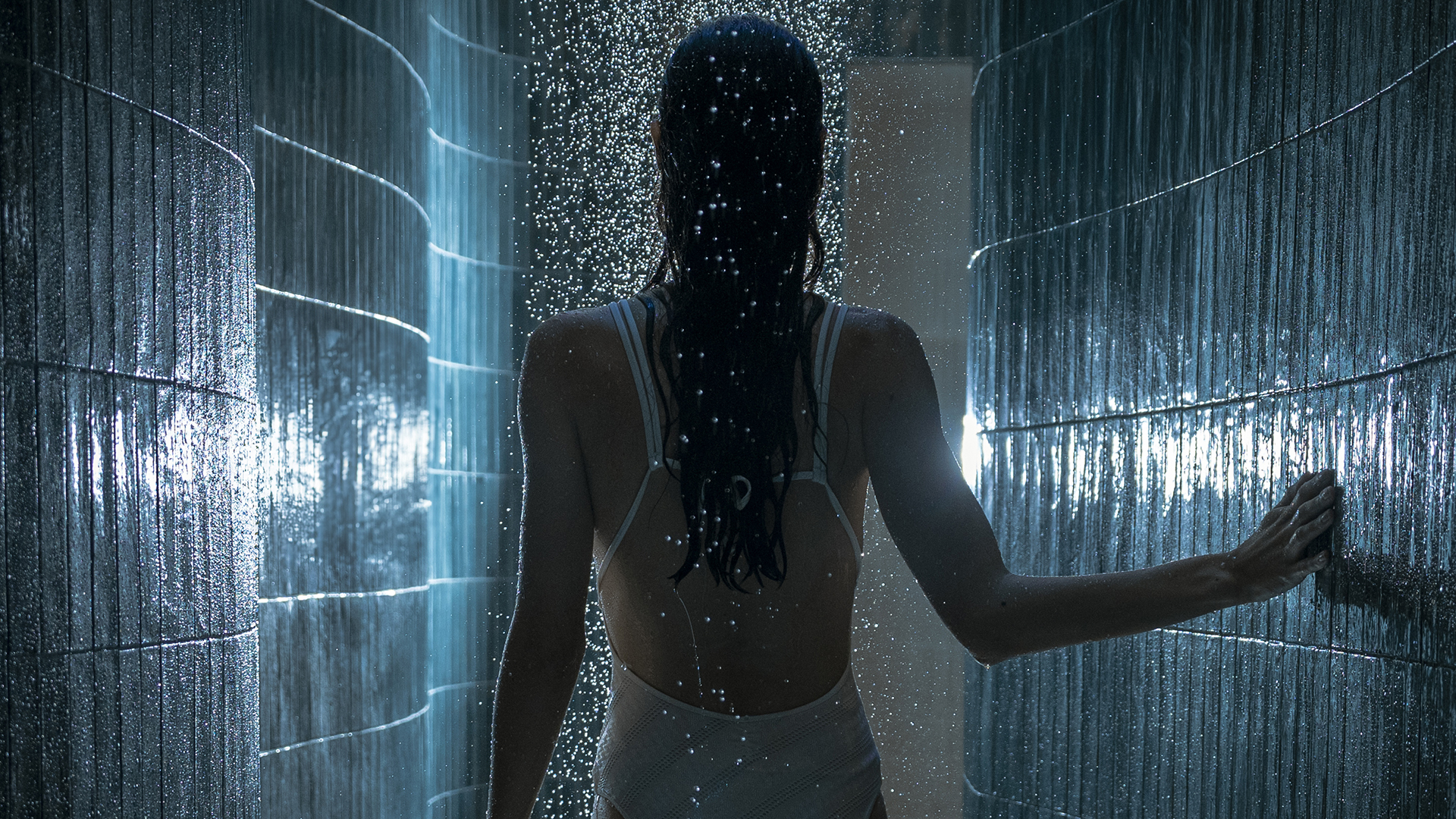Woman walking through a rainforest-style hydrotherapy shower, surrounded by illuminated water streams and textured blue walls.