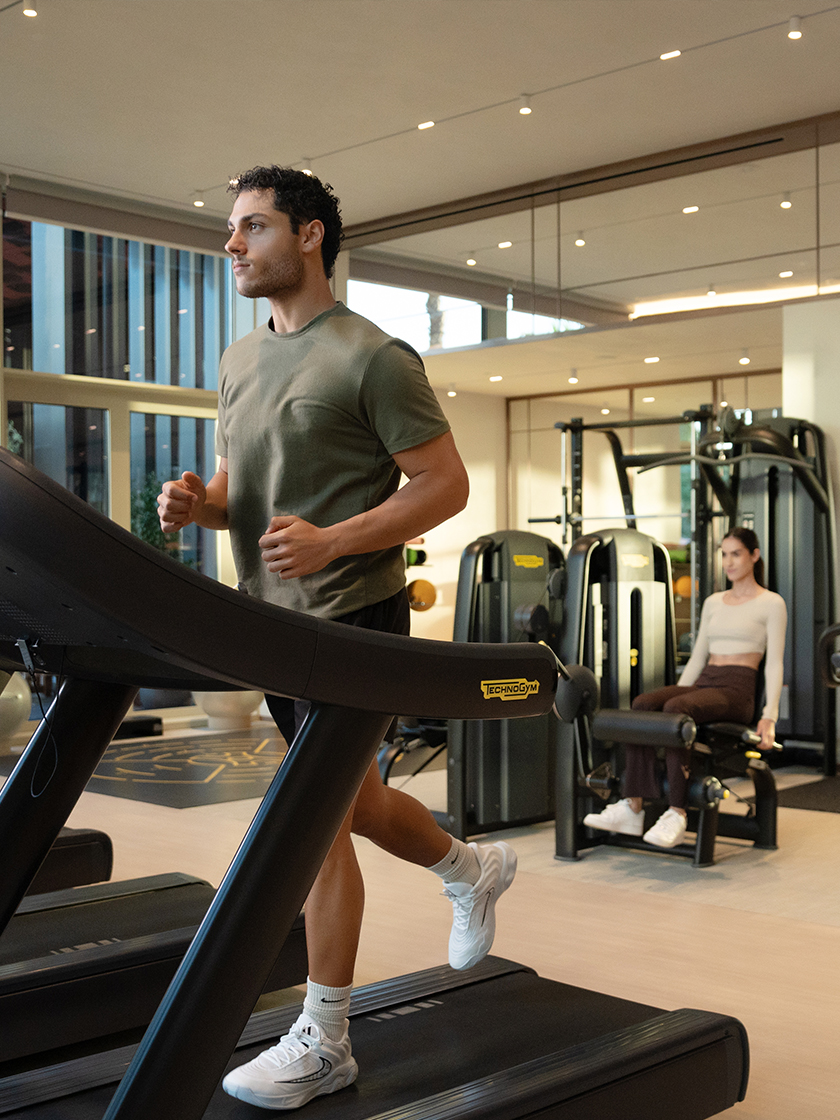 Man running on a treadmill in a modern gym while a woman trains on strength equipment in the background.