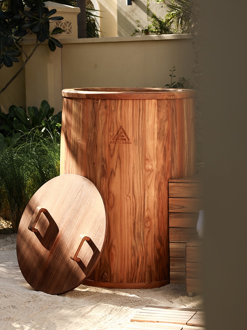 Wooden Arctic Edge cold plunge tub placed outdoors in a serene garden setting, with its round lid resting nearby.