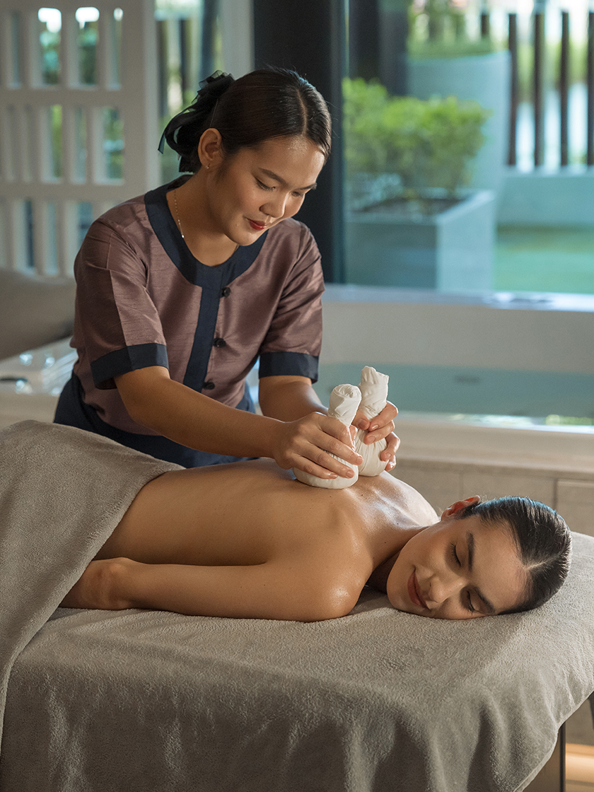Spa therapist performing a warm herbal compress massage on a guest’s back in a serene treatment room.