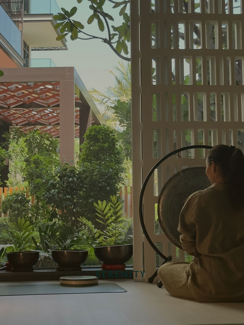 Wellness practitioner seated indoors playing a large gong, with singing bowls and lush garden views creating a serene meditation space.