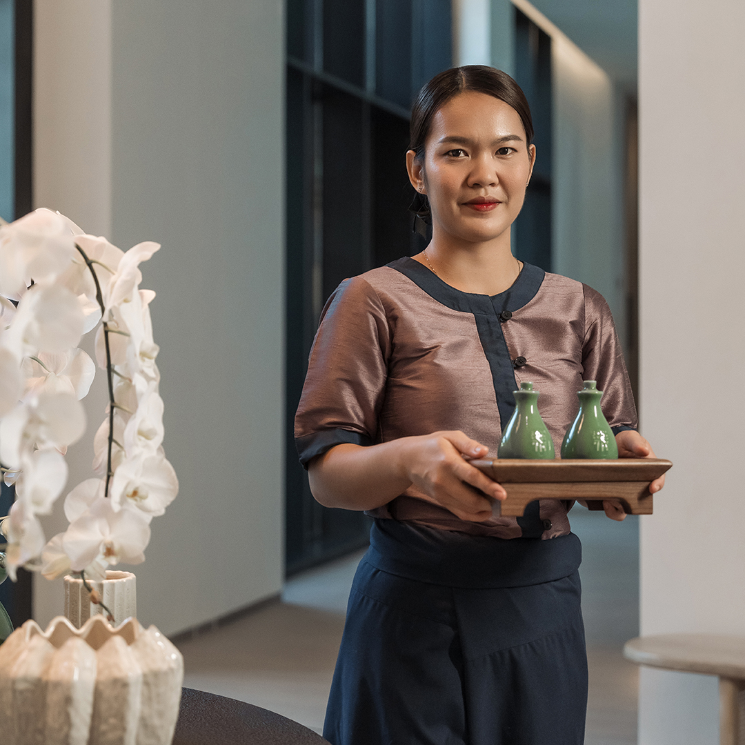 Spa therapist warmly welcoming guests while holding a tray with ceramic wellness oils in a serene reception area.