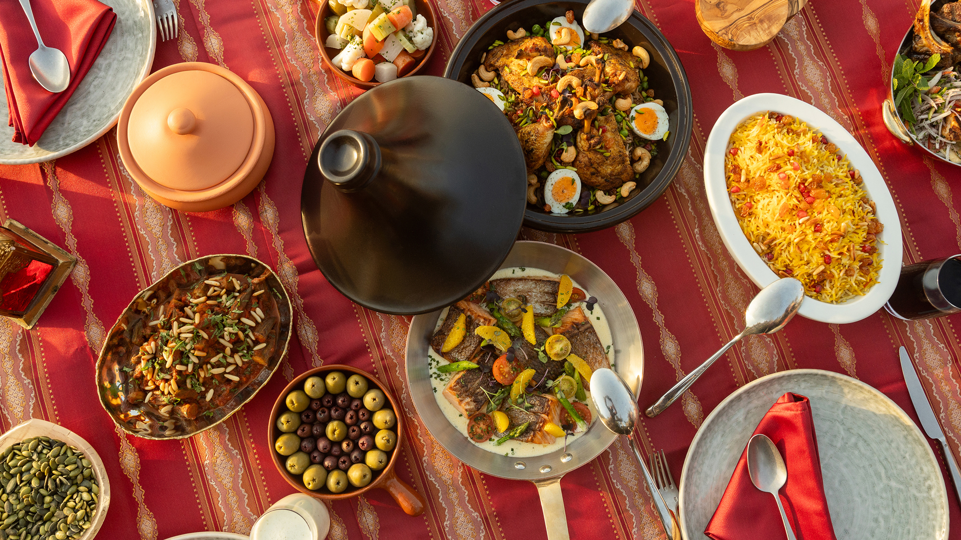 Overhead view of a beautifully arranged Middle Eastern dining table featuring a variety of colourful dishes, including rice, grilled meats, vegetables, olives and shared plates on a red patterned tablecloth.