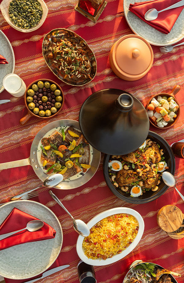 Overhead view of a beautifully arranged Middle Eastern dining table featuring a variety of colourful dishes, including rice, grilled meats, vegetables, olives and shared plates on a red patterned tablecloth.
