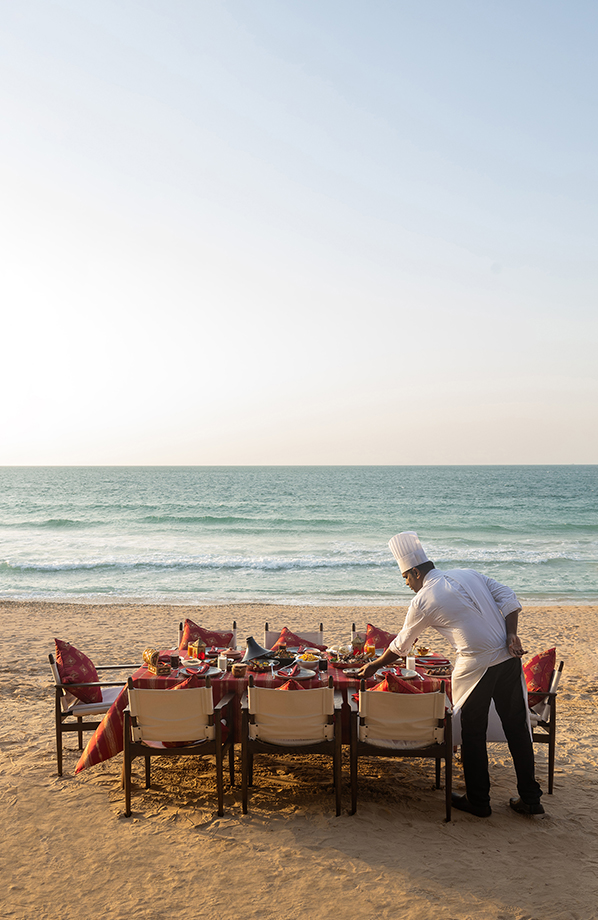 A beachfront dining setup with a beautifully set table and a chef preparing dishes beside the ocean at sunset, creating an intimate seaside dining experience.