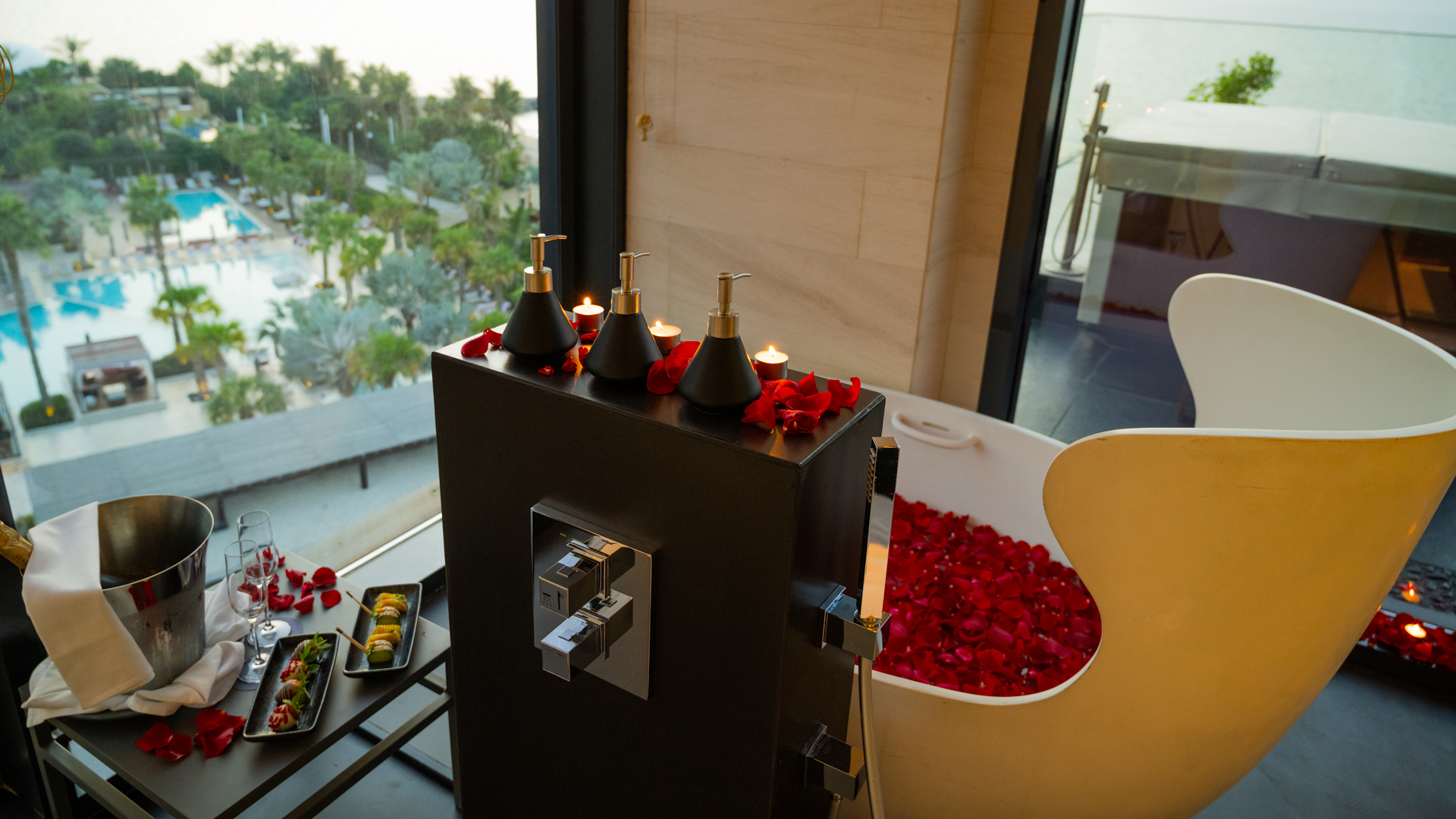 Romantic in-room bathtub filled with rose petals beside lit candles and bath amenities, overlooking the resort pool and beachfront at sunset.