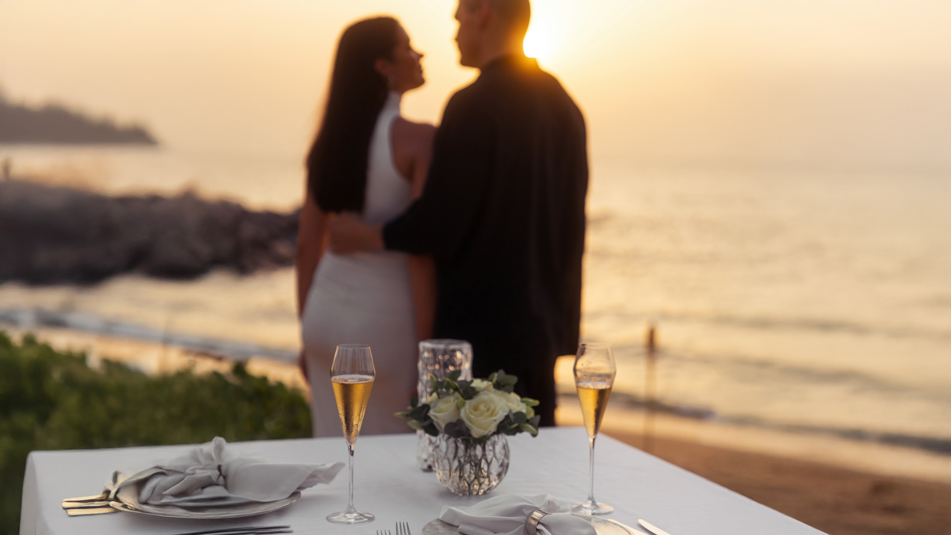 Couple embracing at sunset on a private beach, with a romantic table set for two featuring champagne glasses and floral décor overlooking the sea.
